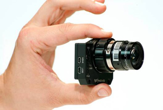 A person's hand holds a small, rectangular black camera module with a lens and various ports.