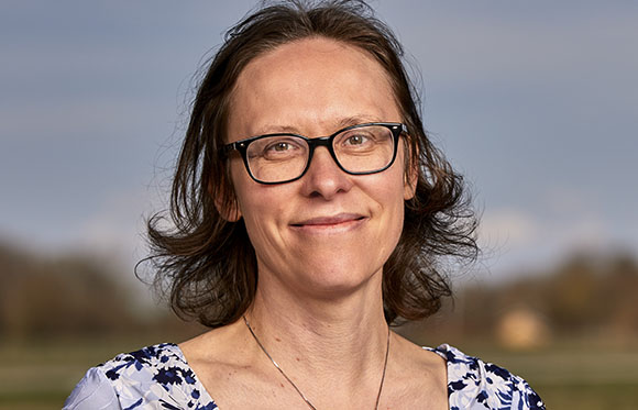 A woman with brown hair and glasses smiling against a soft-focus outdoor background.