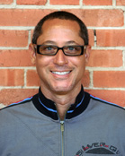 A smiling man with short dark hair and glasses wearing a grey zip-up jacket in front of a brick wall.
