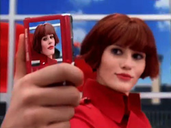 A woman with short red hair wearing a red coat poses for a selfie.