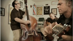 Three men playing a double bass, an acoustic guitar, and a mandolin in a room with framed art.