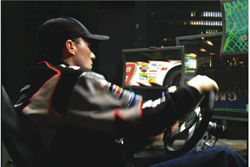 A man in a cap and racing suit sits in a chair, holding a steering wheel while looking at racing simulator screens.