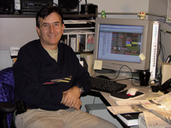 A man in a dark sweater sits at a desk with a computer monitor, keyboard, and mouse.