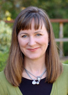 A woman with shoulder-length brown hair and bangs smiles at the camera outdoors.