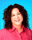 A smiling woman with dark curly hair wearing a pink collared shirt against a bright blue background.
