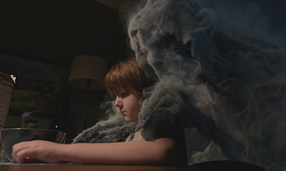 A young person sits at a table partially obscured by thick, swirling grey smoke in a dimly lit room.