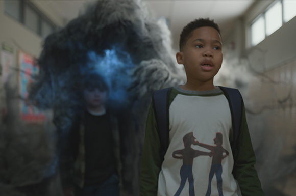 A young boy in a graphic t-shirt looks ahead in a hallway with a large, dark smoke cloud behind him.