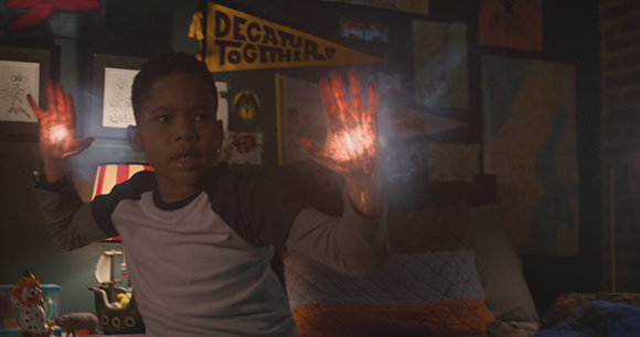 A young boy holds up both hands, which are glowing with bright, magical light in a dark bedroom.
