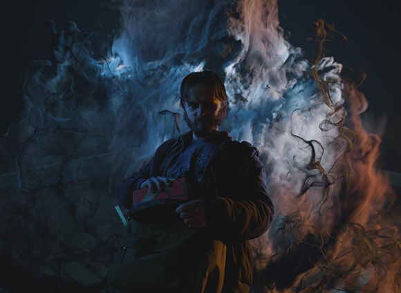 A man with a beard stands centered behind thick, swirling clouds of blue and orange smoke.