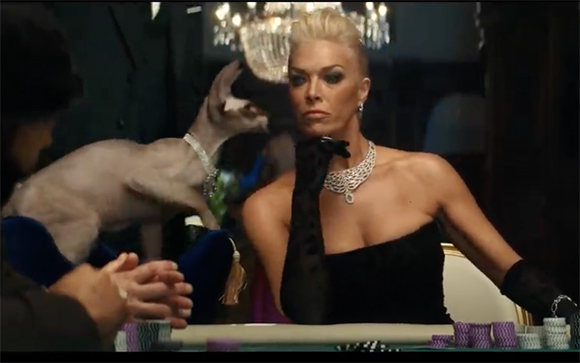 A blonde woman in a black strapless dress and jewelry sits at a table as a small animal reaches toward her.