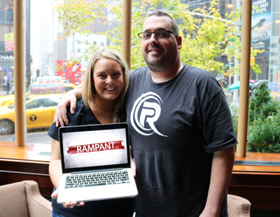 A man and woman smile while holding a laptop that shows the word RAMPANT in red text on a white background.