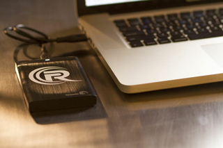 A black external hard drive with a circular logo connected via USB to a silver laptop on a desk.