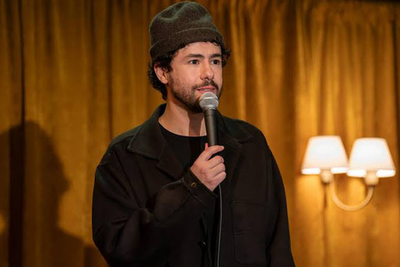 A man with a beard and beanie holds a microphone while speaking in front of gold curtains.