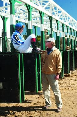 A jockey in blue and white silks sits on a green starting gate next to a man in a tan jacket.