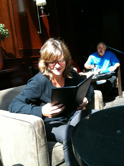 A woman in a black sweater sits in an armchair reading a black menu while a man sits at a table behind her.