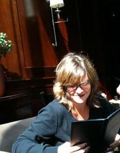 A woman with blonde hair and glasses reads a black book in a dimly lit room.