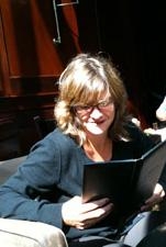 A woman with blonde hair and glasses smiling while reading a black book.