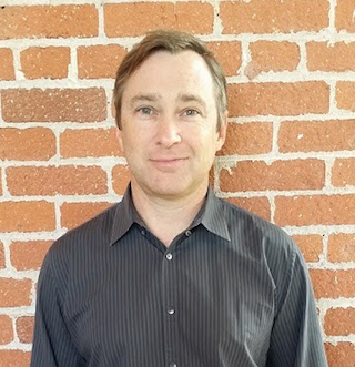 A man with short brown hair wearing a dark grey striped button-down shirt against a red brick wall.