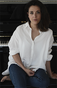 A woman with dark hair wearing a white button-down shirt and blue jeans sits in front of a black piano.