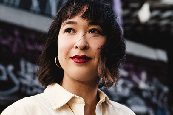 A woman with dark hair and red lipstick looks upward against a blurred urban background.