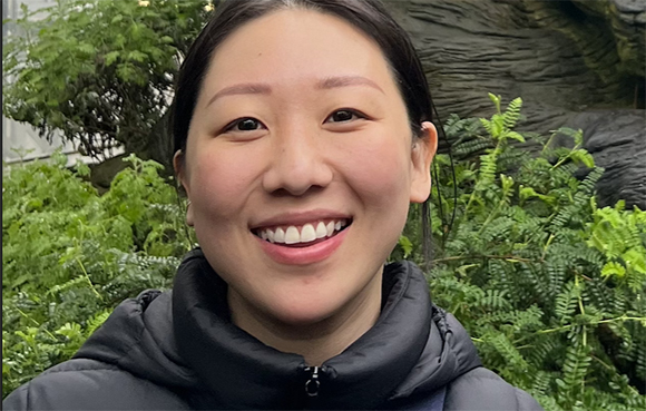 A smiling woman with dark hair wearing a black puffer jacket in front of green foliage.