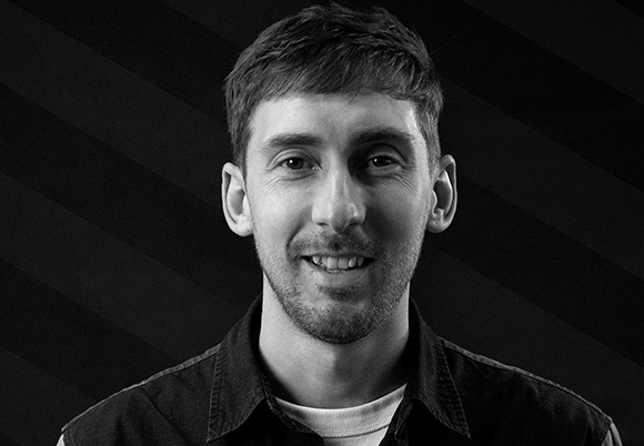 Black and white headshot of a smiling man with short hair and a beard against a dark striped background.