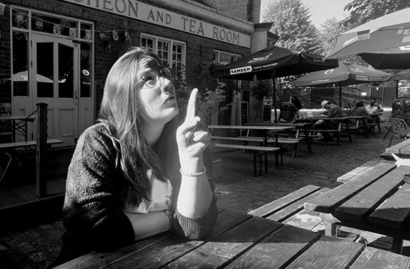 A woman with glasses looks upward while gesturing with one finger at an outdoor cafe seating area.