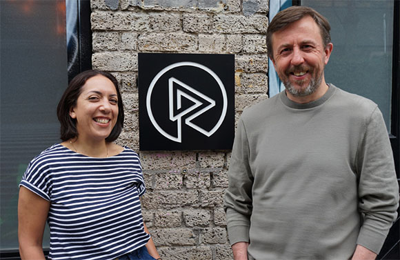 A woman in a striped shirt and a man in a grey sweatshirt stand beside a circular logo on a brick wall.