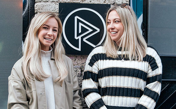 Two smiling blonde women standing in front of a circular black logo on a brick wall.