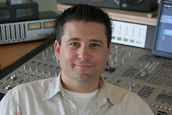 A man smiling in front of a large professional audio mixing console in a studio.