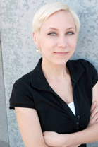 A blonde woman with short hair and a black polo shirt smiles against a grey textured wall.