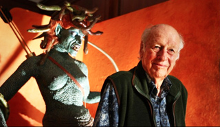 An elderly man smiling next to a large, blue-skinned Medusa statue with snake hair against an orange background.