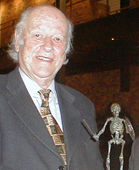 Smiling elderly man in a dark suit and patterned tie standing next to a small skeleton figurine.