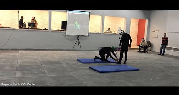 A person in a black suit kneels on a blue mat in a large studio with a projection screen in the background.