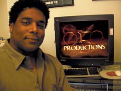 A man in a tan shirt sits next to a computer monitor displaying the text 'PRODUCTIONS' with a gold logo.