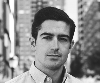 Black and white headshot of a man with dark hair looking directly at the camera against a city background.