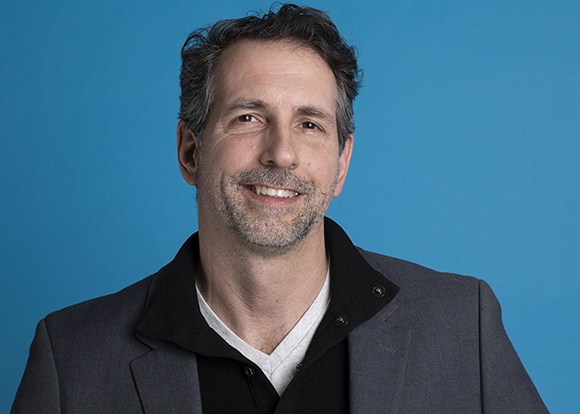 A smiling man with salt-and-pepper hair wearing a grey blazer and black shirt against a blue background.