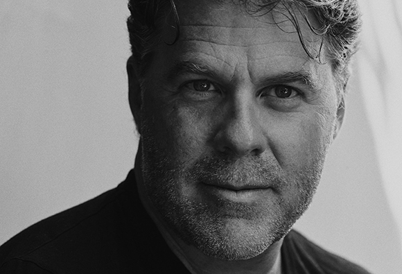 Black and white close-up portrait of a man with short, wavy hair and light stubble looking at the camera.