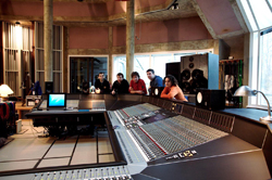 A group of people gathered behind a large mixing console in a recording studio.