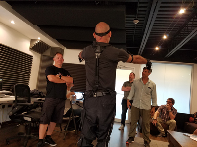 A man in a black motion capture suit and headset stands with arms outstretched in a studio.