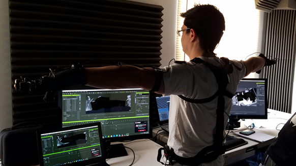 A person in a white shirt and motion capture harness extends their arms toward computer monitors.