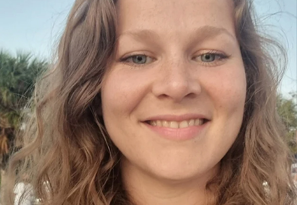A smiling woman with wavy brown hair and blue eyes outdoors.