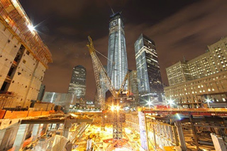 A construction site at night with cranes and illuminated skyscrapers under a dark sky.