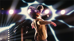 A person in a tan suit performs a dance move under bright, radiating stage lights.