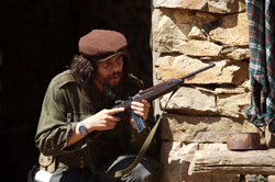 A man with long hair and a beard wearing a brown cap and olive jacket holds a rifle near a stone wall.