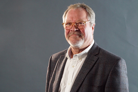 A man with graying hair, glasses, and a mustache wearing a white shirt and a dark blazer.