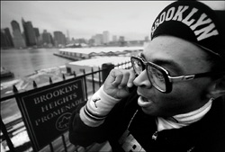 Black and white close-up of a man in a Brooklyn cap talking on a phone by a waterfront city skyline.