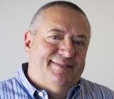 A smiling man with short graying hair wearing a blue and white striped button-down shirt.
