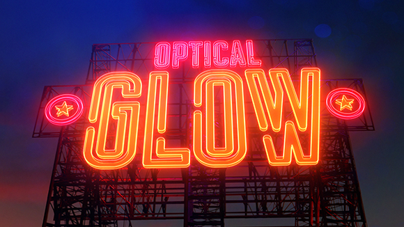 A large, bright orange and red neon sign reading 'OPTICAL GLOW' mounted on a dark metal scaffolding structure.