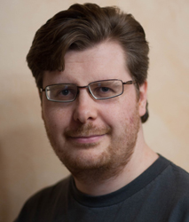 A man with brown hair and glasses smiling slightly against a neutral background.